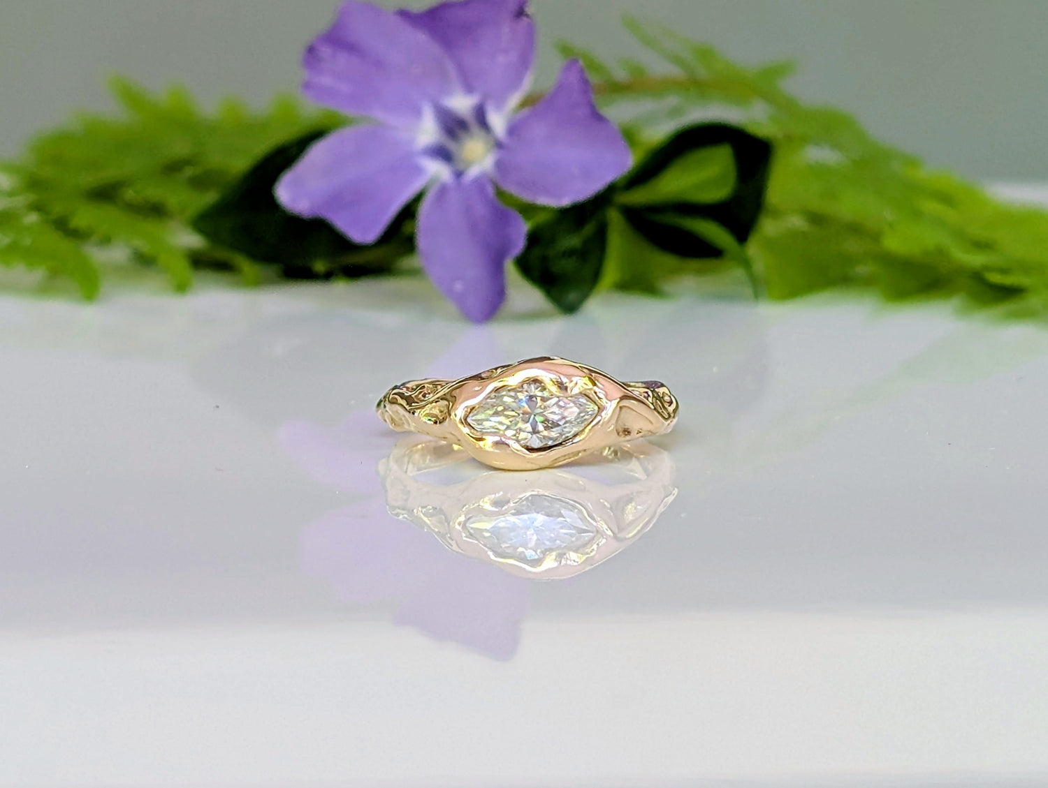 Gold ring with a diamond on a reflective surface with a purple flower and green leaves in the background.