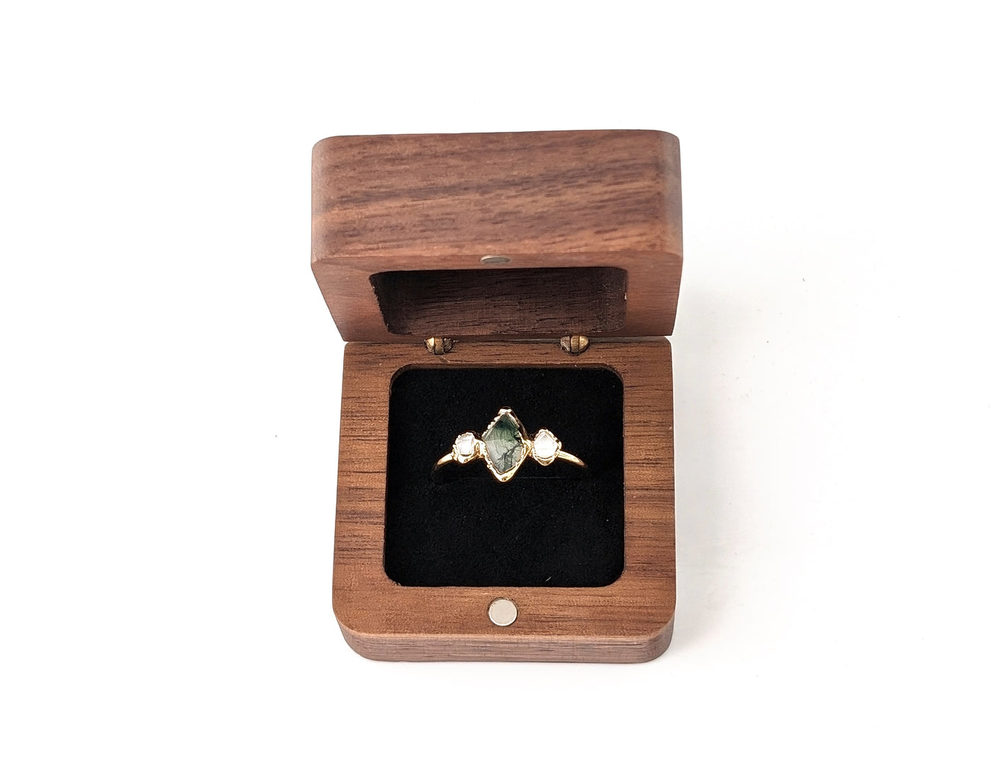 Square wooden ring box open with a green gemstone ring and two pearls inside on a black velvet lining.