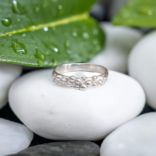 Dew Drop textured silver ring showcasing beaded design on smooth pebbles with green leaves in background.
