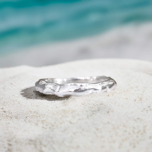 Molten Silver wedding band resting on sandy beach, showcasing its unique irregular and handcrafted design.