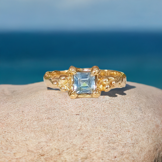 London Blue Topaz engagement ring in solid 14k gold with an organic textured band, set against a serene ocean backdrop.