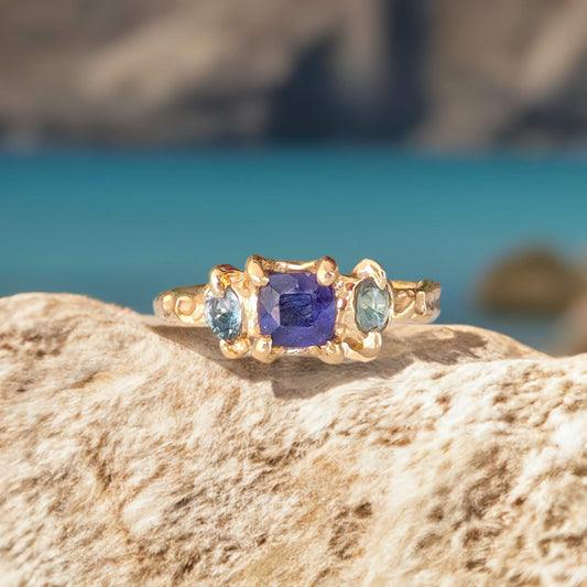 Elegant Blue Sapphire and Tourmaline engagement ring in solid 14k gold on a rocky surface with a beach background.