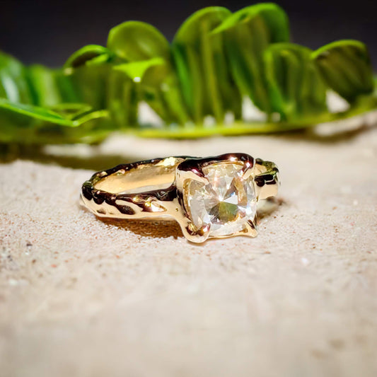 Gold Moissanite textured engagement ring with an 8mm cushion cut stone, set on sandy background with greenery.