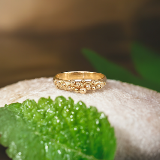 Solid gold beaded Dew Drop ring on a soft surface with green leaves, showcasing its unique molten design.