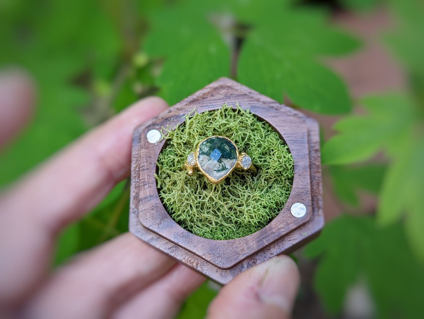 Pear-shaped moss agate and raw diamond ring displayed on a wooden hexagon with green moss background.