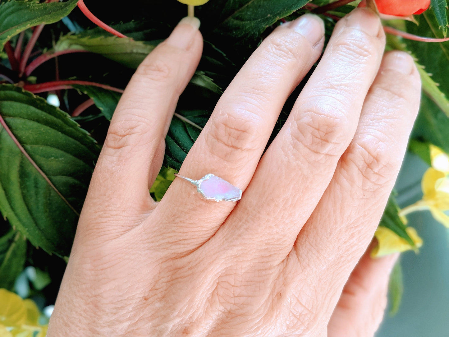 Hand wearing a rustic raw pink Morganite ring on a delicate Sterling Silver band, surrounded by green foliage.