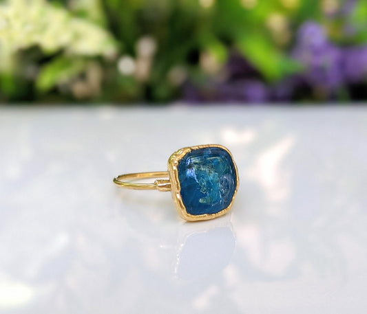Natural blue Apatite ring on a gold band, showcasing a unique stone design with a blurred floral background.