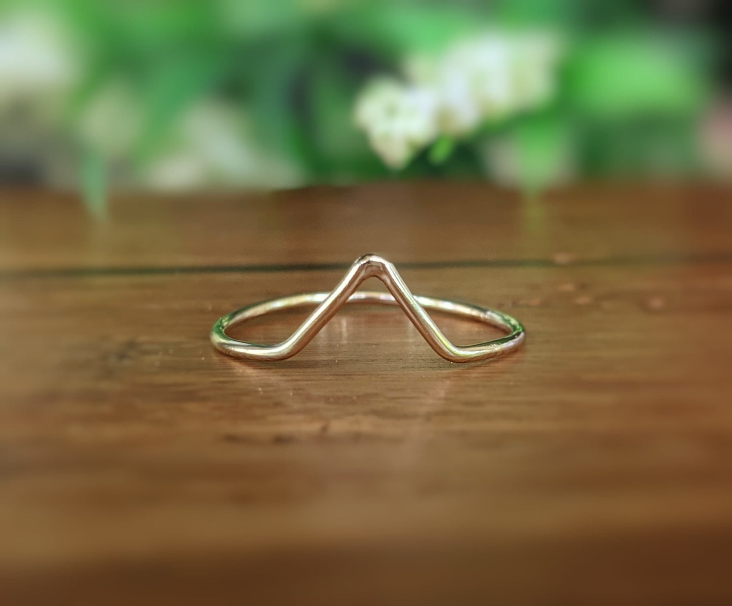 18k gold chevron band ring displayed on wooden surface with blurred floral background.