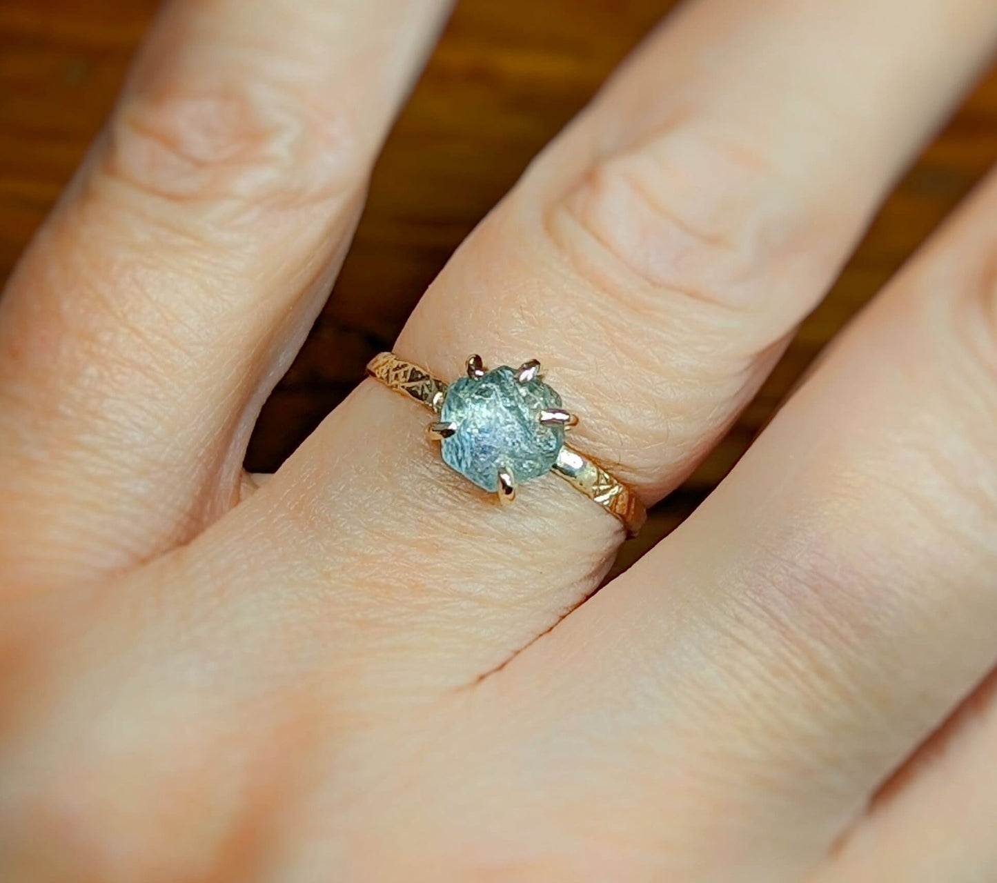 Teal-blue raw Montana Sapphire engagement ring on a hand with a textured gold band, symbolizing love and devotion.