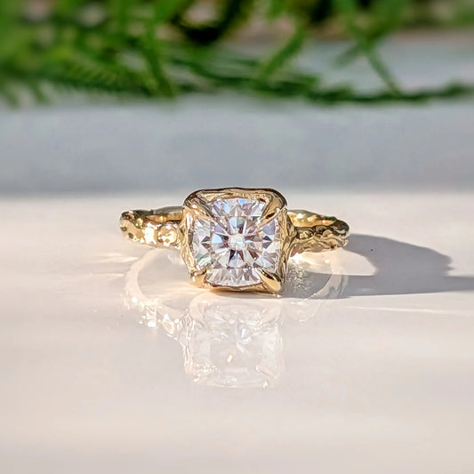 A gold engagement ring with a large, clear diamond set in the center, surrounded by a decorative band, is displayed on a white surface with a blurred green plant in the background.