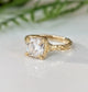 A close-up image of a gold engagement ring with a large, clear gemstone set in a square-cut diamond.