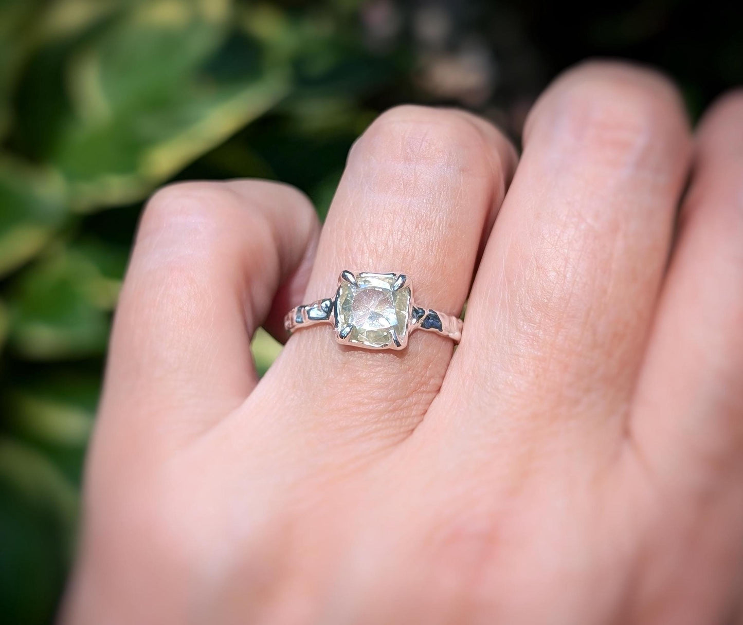 Silver moissanite engagement ring with cushion cut stone displayed on a hand against a blurred green background.