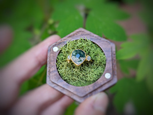 Pear shape Moss Agate ring with raw diamond accents, set on a wooden hexagonal display with moss.