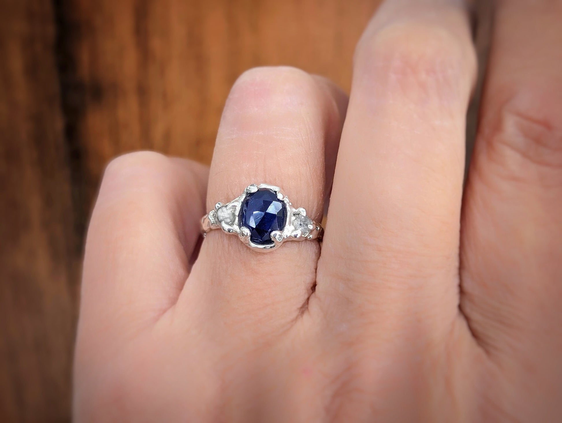Woman's hand wearing an Oval Blue Sapphire and 2 small raw diamonds set by prongs on a textured Molten Solid Sterling Silver band